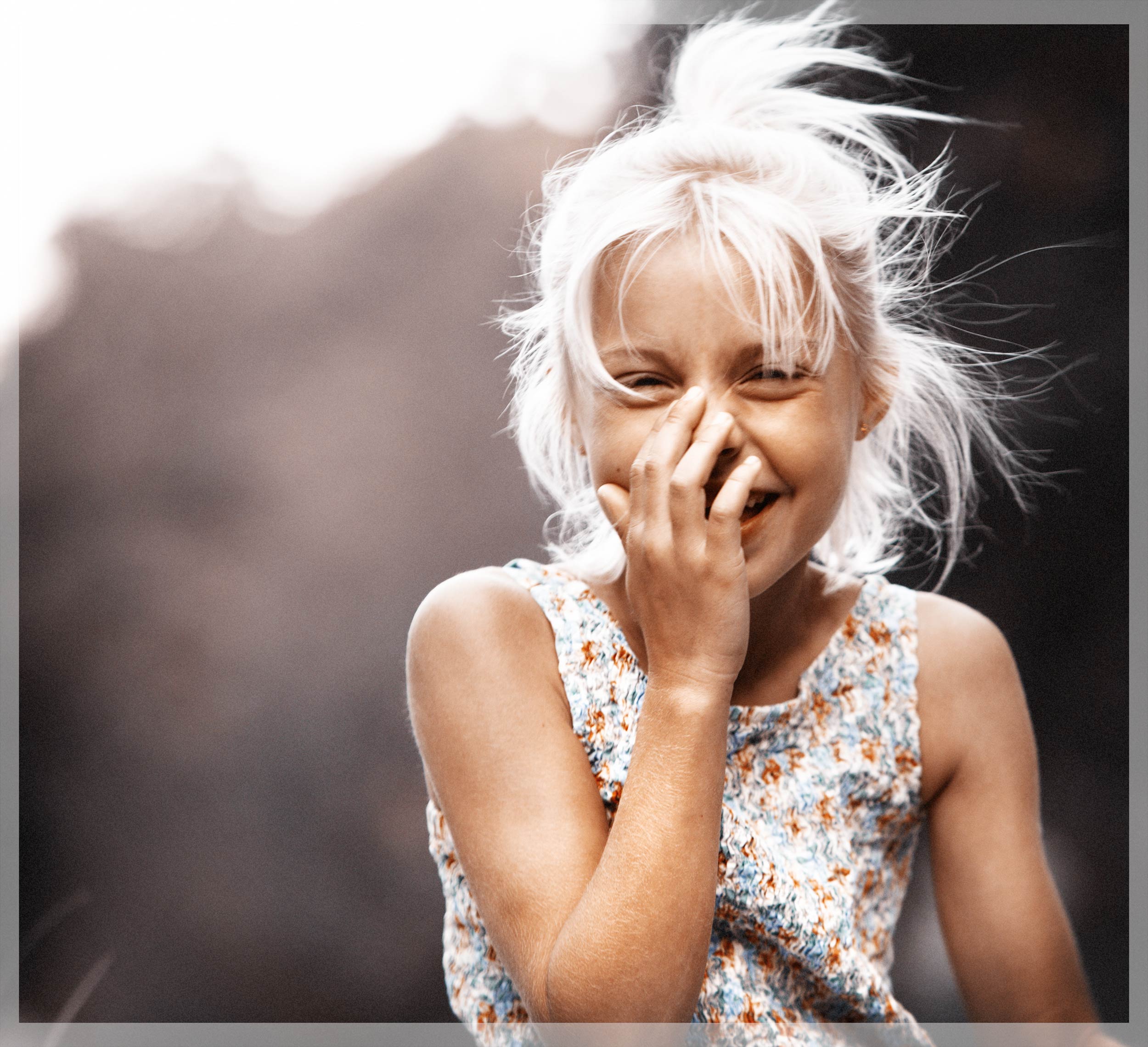 A young blonde girl covering her mouth as she laughs with the wind blowing her hair